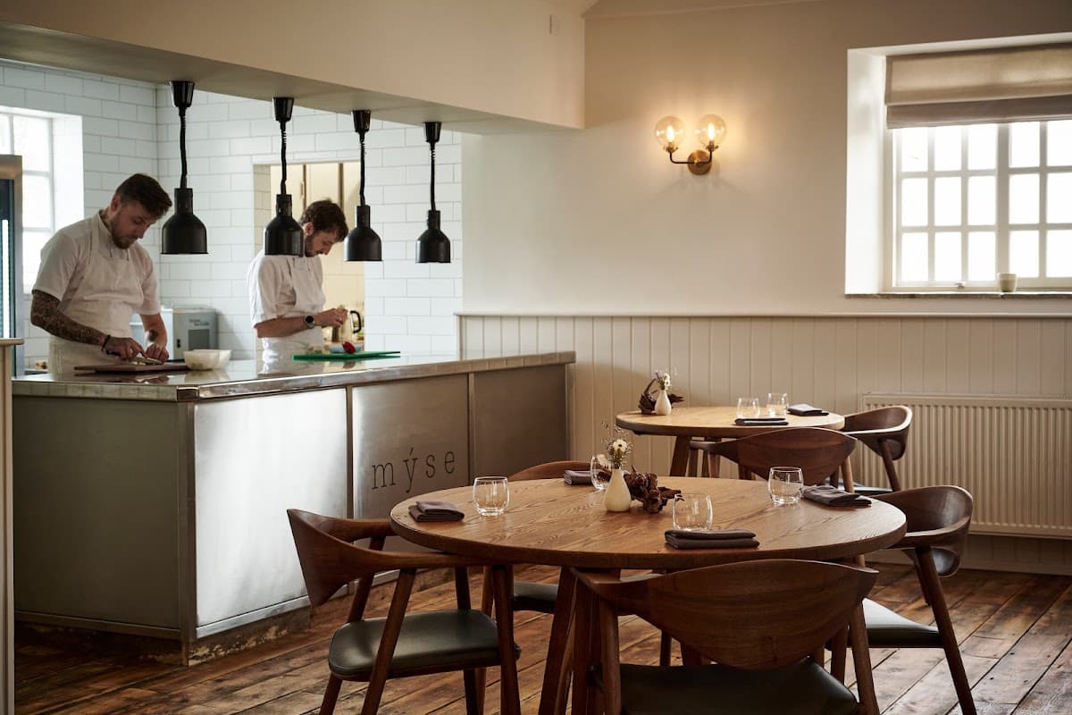 Modern restaurant interior with wooden tables, stylish lighting, and chefs preparing food in an open kitchen.